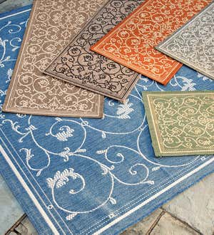 Several Veranda Scroll indoor/outdoor rugs in different colors layered on a stone patio.