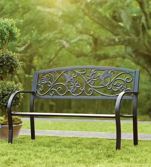 METAL FURNITURE > A metal garden bench with a decorative scroll and hummingbird motif in a yard. 