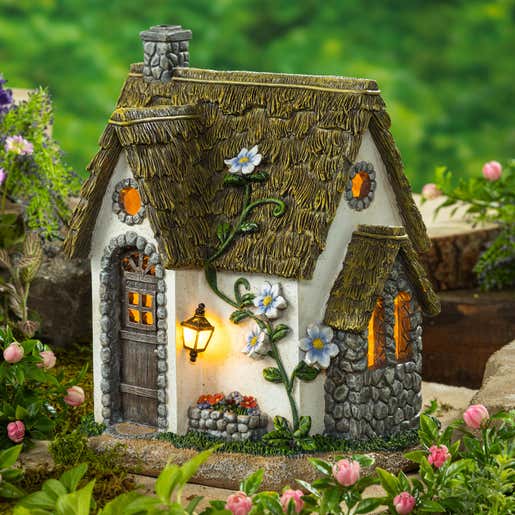 A miniature fairy cottage with glowing windows and lantern sits in a garden bed