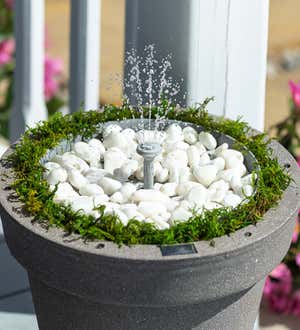 A bubbling water Hydria fountain kit installed in a green planter in an outdoor flower garden.