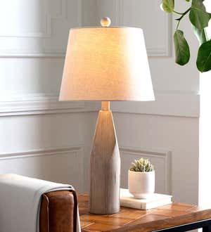 A table lamp with a cream shade and tapered driftwood base next to a sofa.
