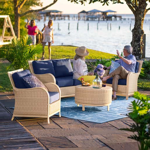 SHOP OUTDOOR SEATING SETS. Image of a couple sitting on a cream Urbanna wicker seating set with deep blue cushions.